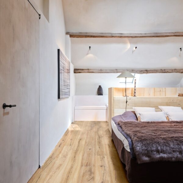 Chambre loft cosy. Plancher bois naturel, poutres apparentes, lit en bois clair et couette marron. Décoration rustique moderne.