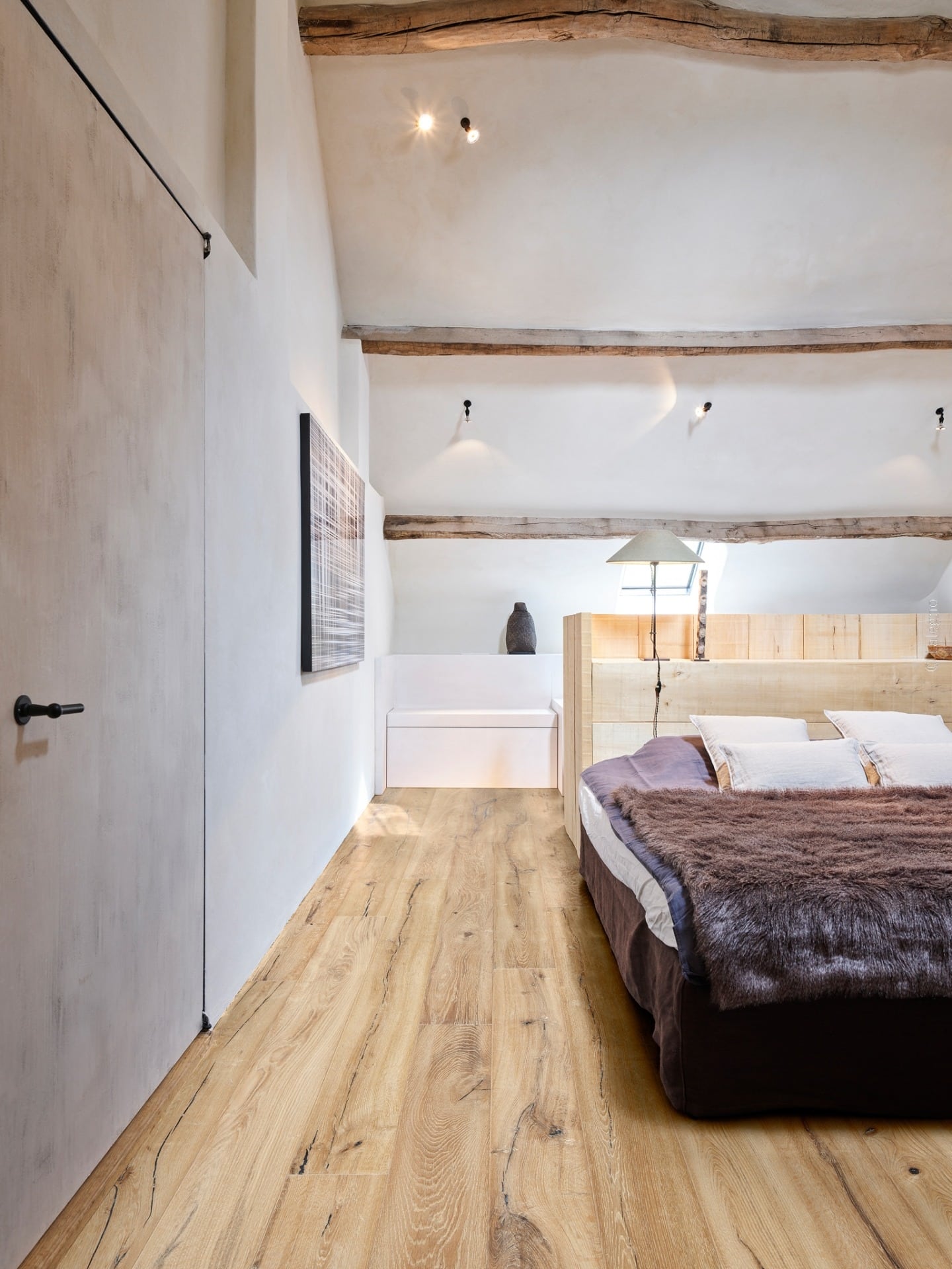 Chambre loft cosy. Plancher bois naturel, poutres apparentes, lit en bois clair et couette marron. Décoration rustique moderne.