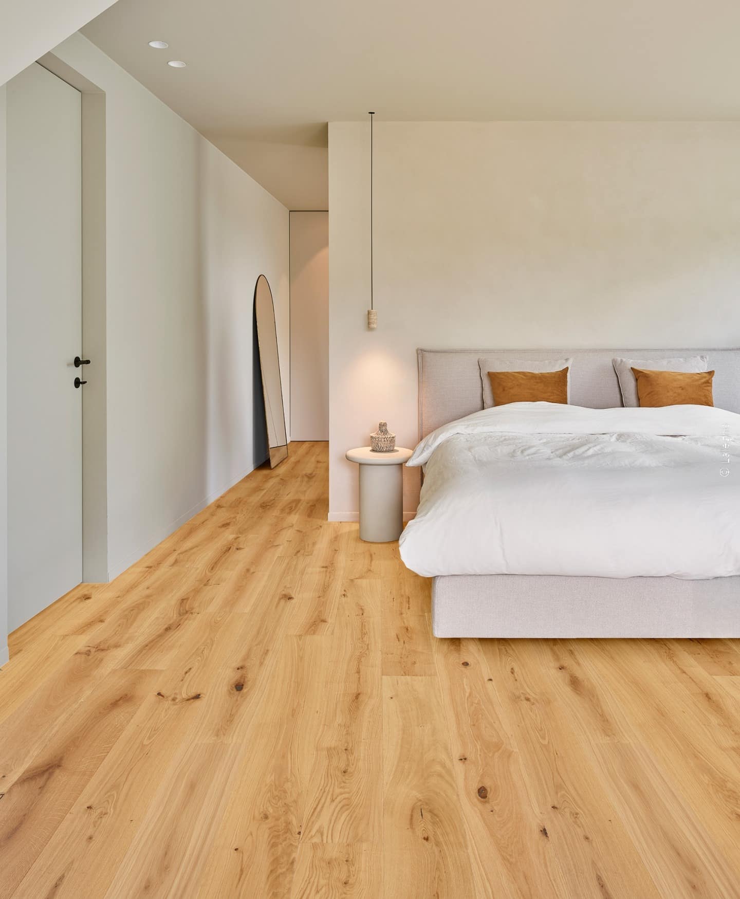 Intérieur de chambre minimaliste: grand lit beige, murs clairs, et parquet en chêne doré. Un couloir mène à un miroir ovale.