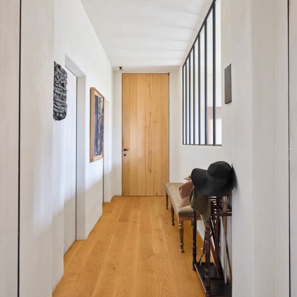 Couloir moderne et minimaliste. Plancher chêne clair, murs blancs et porte en bois naturel. Fenêtre atelier, banc d'entrée et chapeaux.