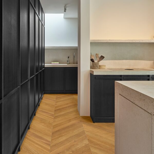 Cuisine moderne: mur de rangement bois noir, sol parquet chêne à chevrons. Îlot et comptoirs en béton clair texturé.
