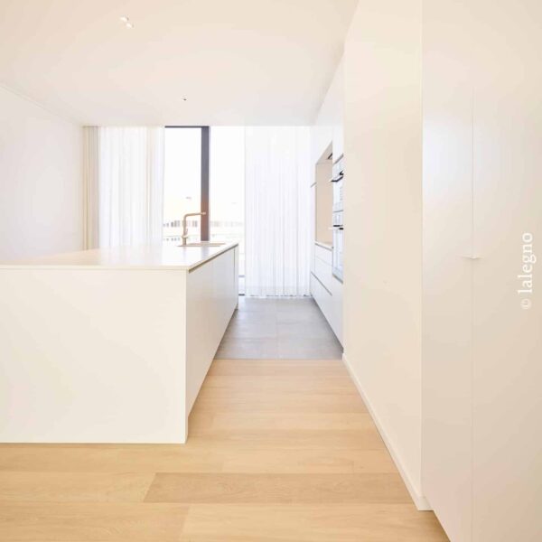 Cuisine minimaliste blanche et lumineuse. Îlot central épuré, robinetterie laiton. Parquet bois clair au premier plan (© lalegno).