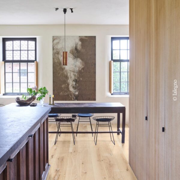 Cuisine rustique moderne, îlot sombre et sol en chêne clair. Longue table devant un tableau abstrait. Panneau mural vertical en bois.