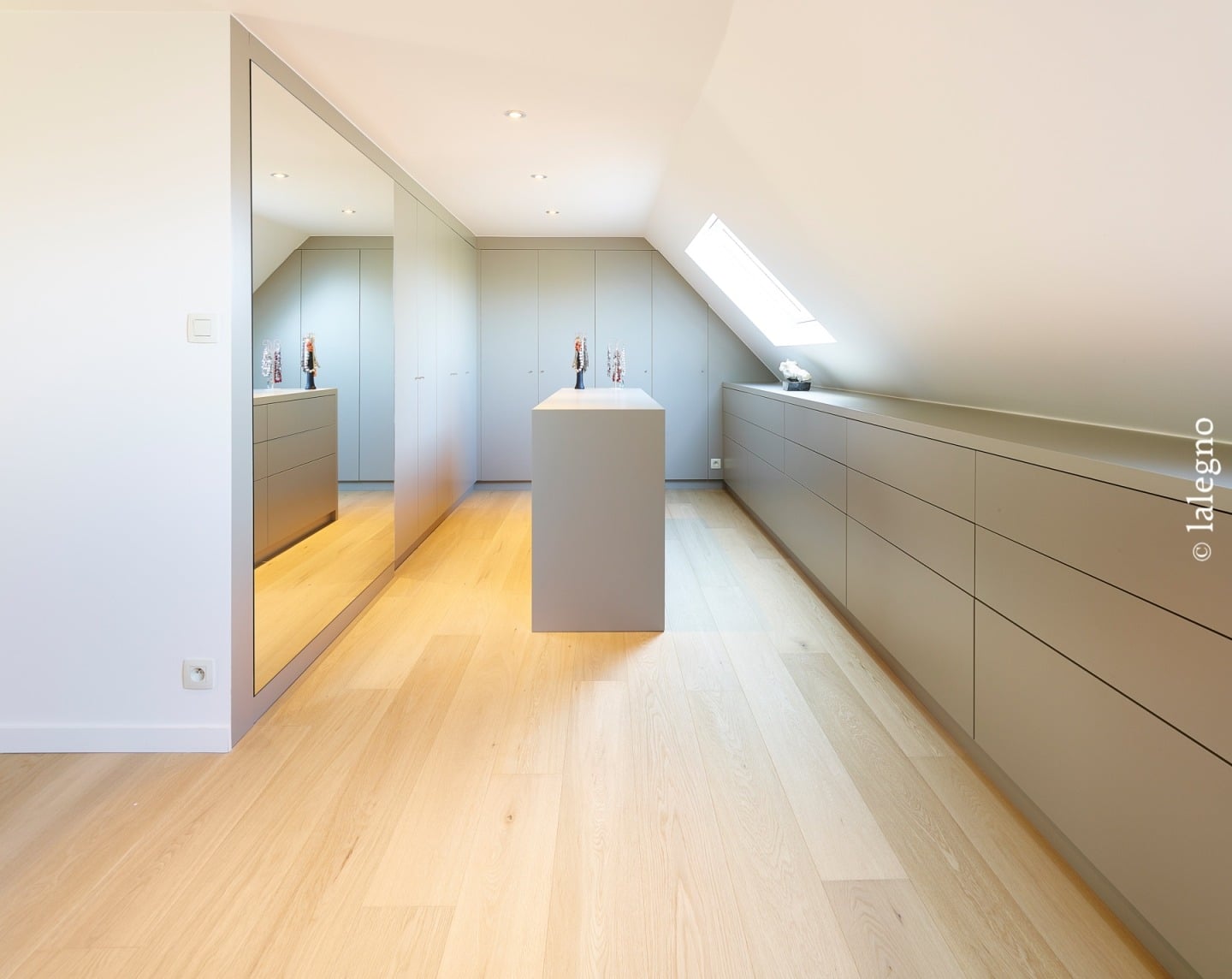 Dressing sous-pente moderne avec armoires, îlot central, et long meuble à tiroirs gris clair. Grand miroir mural et sol en chêne.