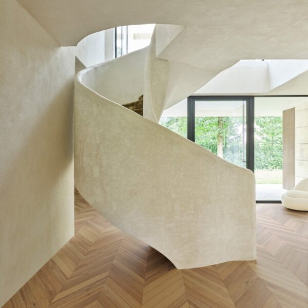 Escalier courbe sculptural en stuc beige texturé dans un hall minimaliste. Parquet chevron bois. Lumière naturelle par porte-fenêtre.