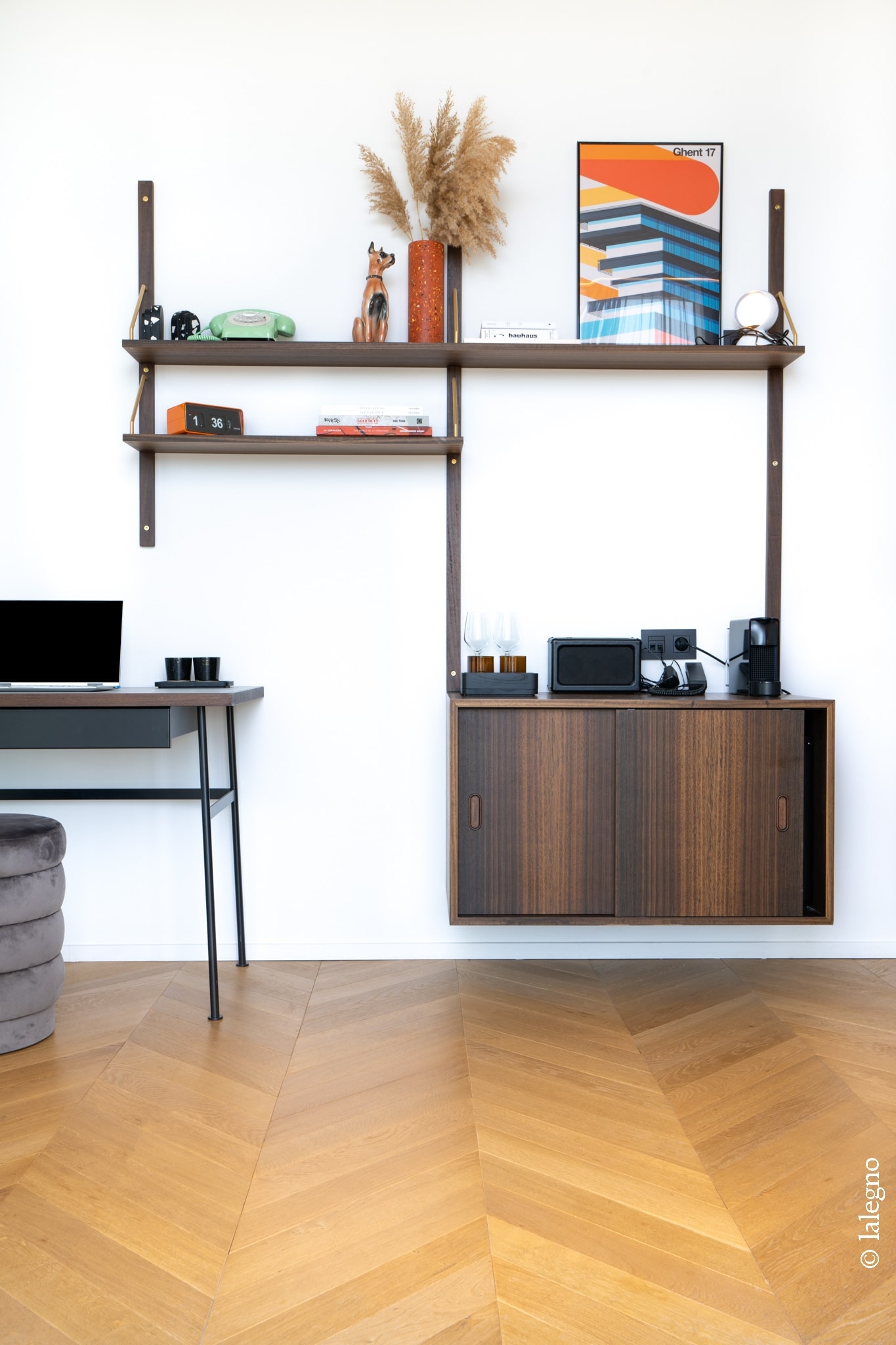 Étagère murale modulaire et meuble en bois foncé dans un bureau. Horloge flip "1:36", poster "Ghent 17". Parquet chêne à chevrons.