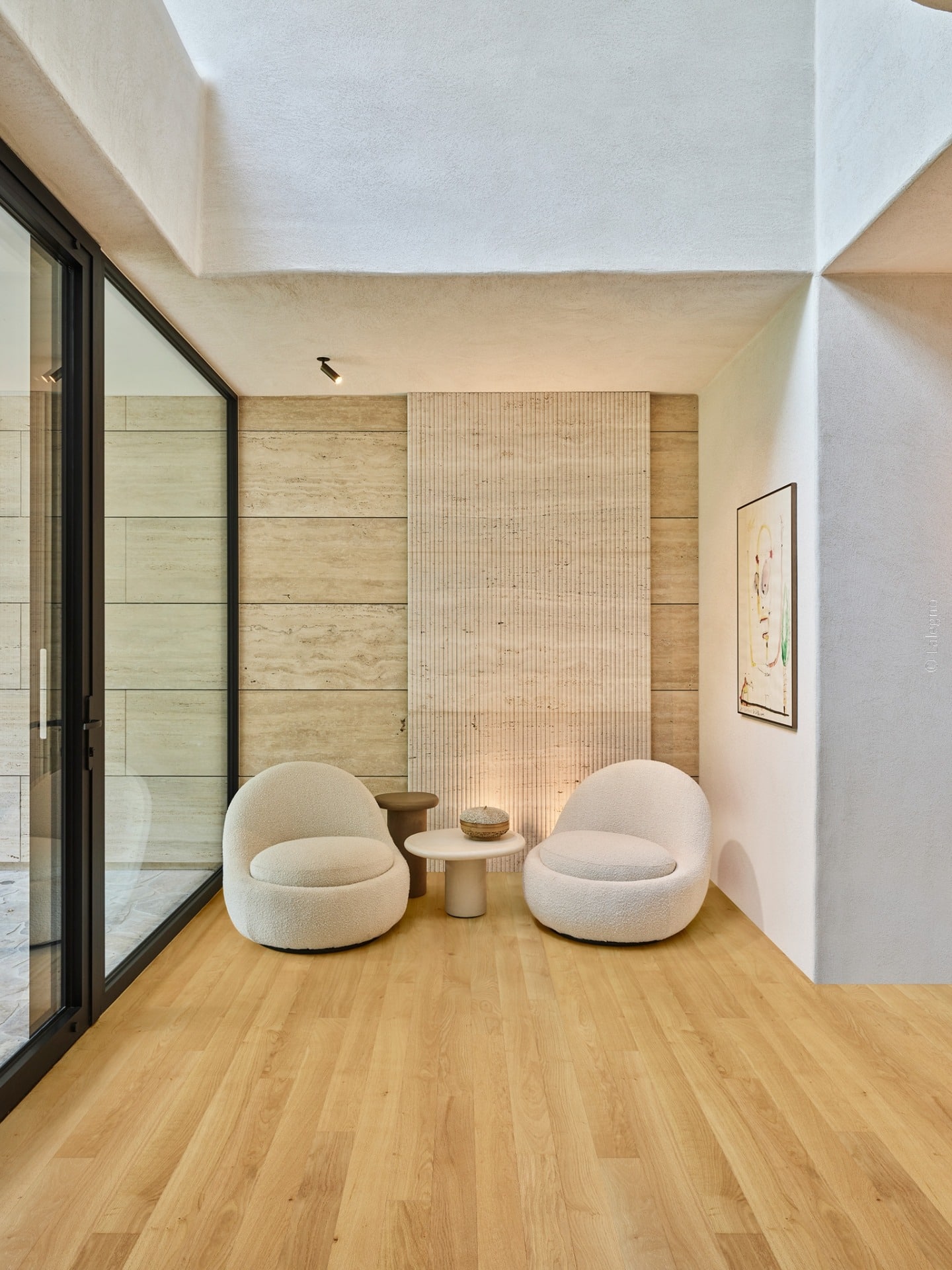 Coin salon moderne: deux fauteuils bouclette moelleux, tables rondes, murs texturés et revêtement en travertin nervuré.