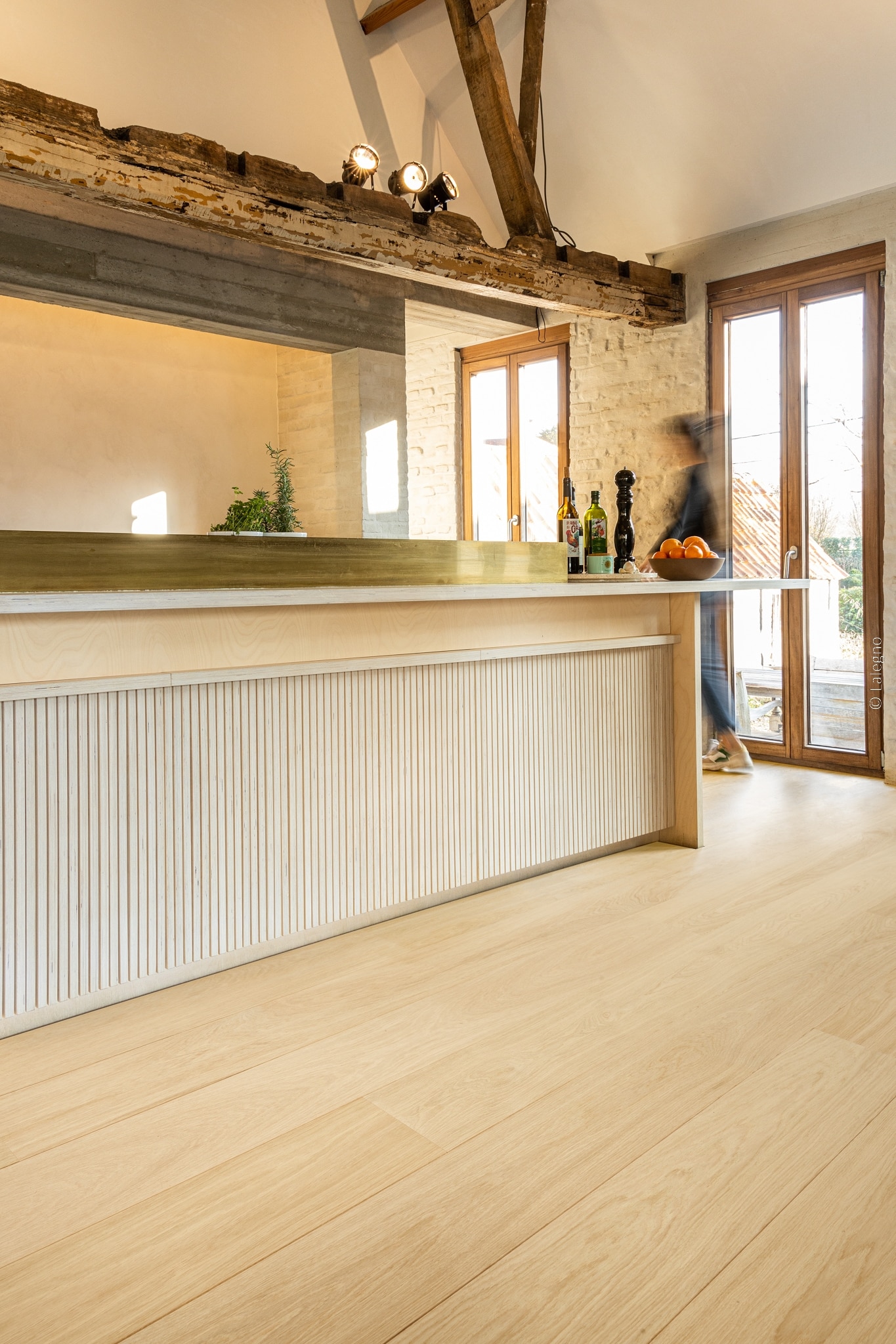 Îlot de cuisine lumineuse en bois clair avec panneau cannelé et laiton. Parquet chêne moderne. Poutres apparentes rustiques en hauteur.