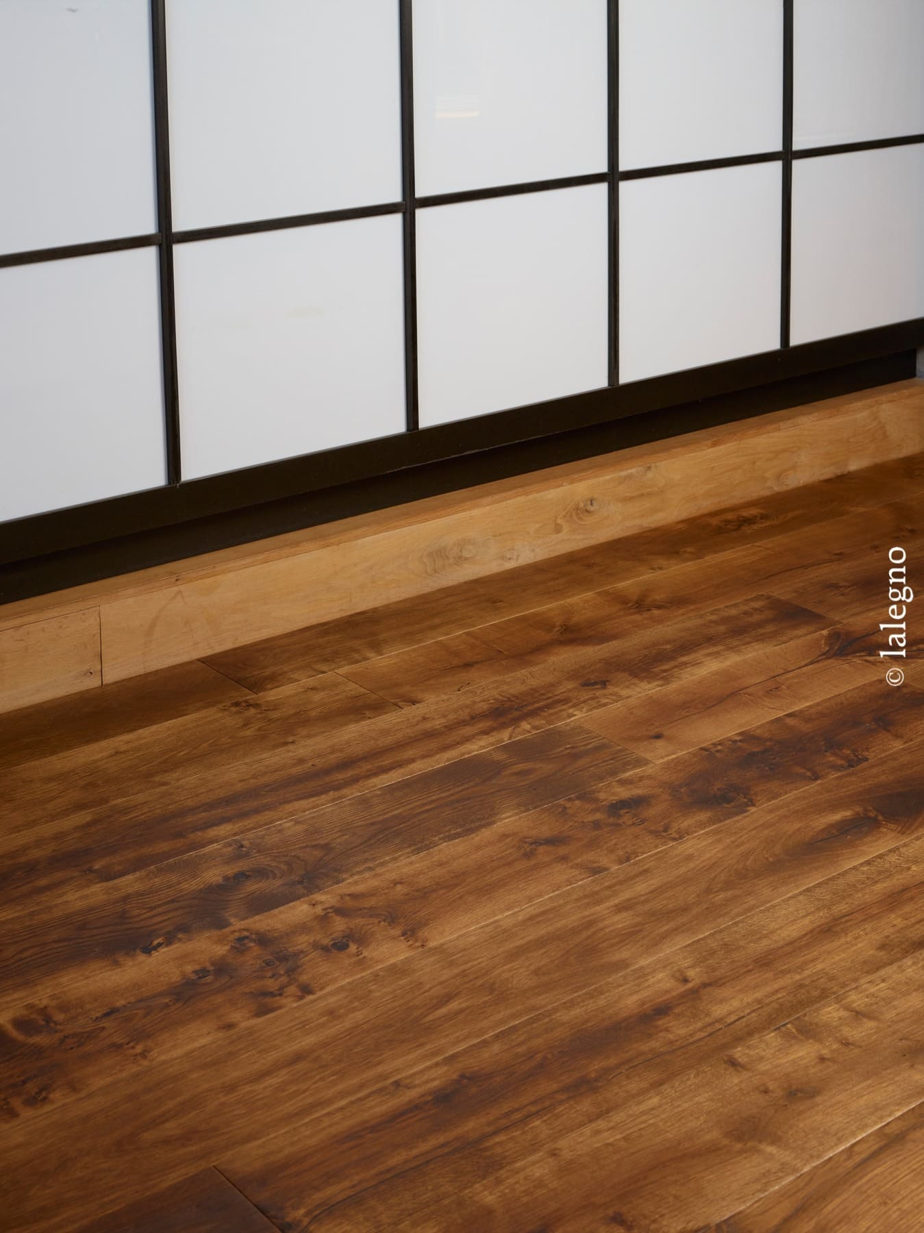 Parquet en bois chêne brun foncé texturé, près d'une plinthe claire. Panneaux muraux blancs quadrillés minimalistes.