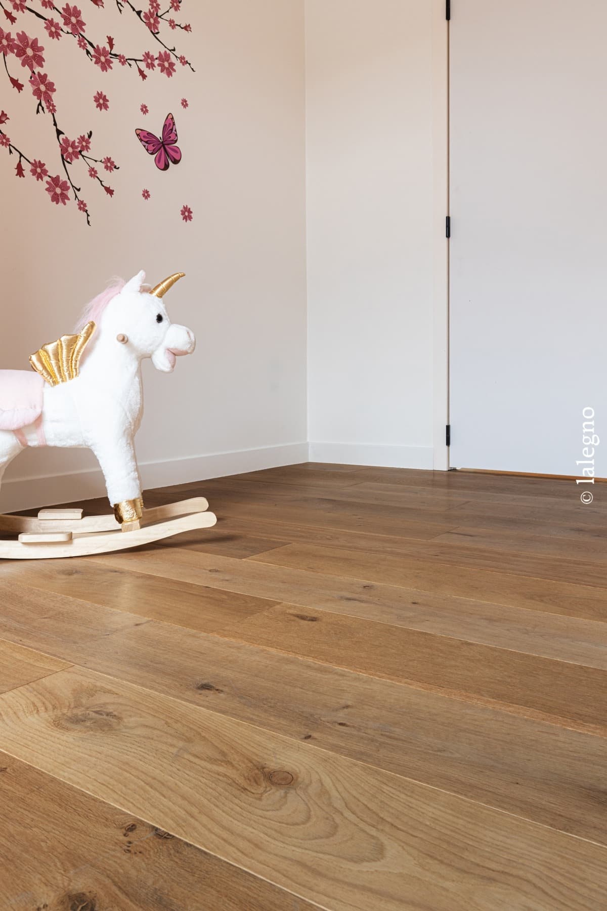 Parquet chêne clair, finition miel, dans une chambre d'enfant. Licorne à bascule blanche et décoration murale fleurie rose.