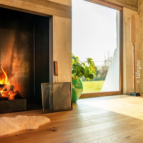 Parquet chêne clair Lalegno devant une cheminée en béton brut, feu chaleureux et grande fenêtre panoramique sur jardin verdoyant.