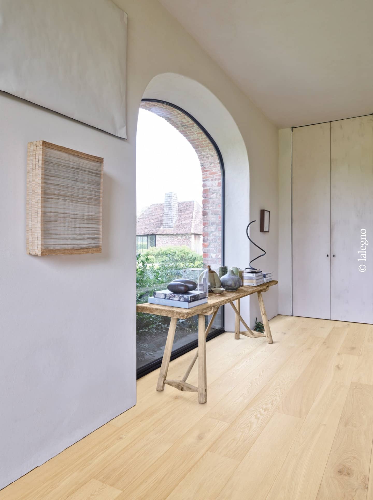 Entrée lumineuse avec large parquet en chêne clair Lalegno. Console rustique, œuvres d'art murales et fenêtre en arc. Style rustique chic.