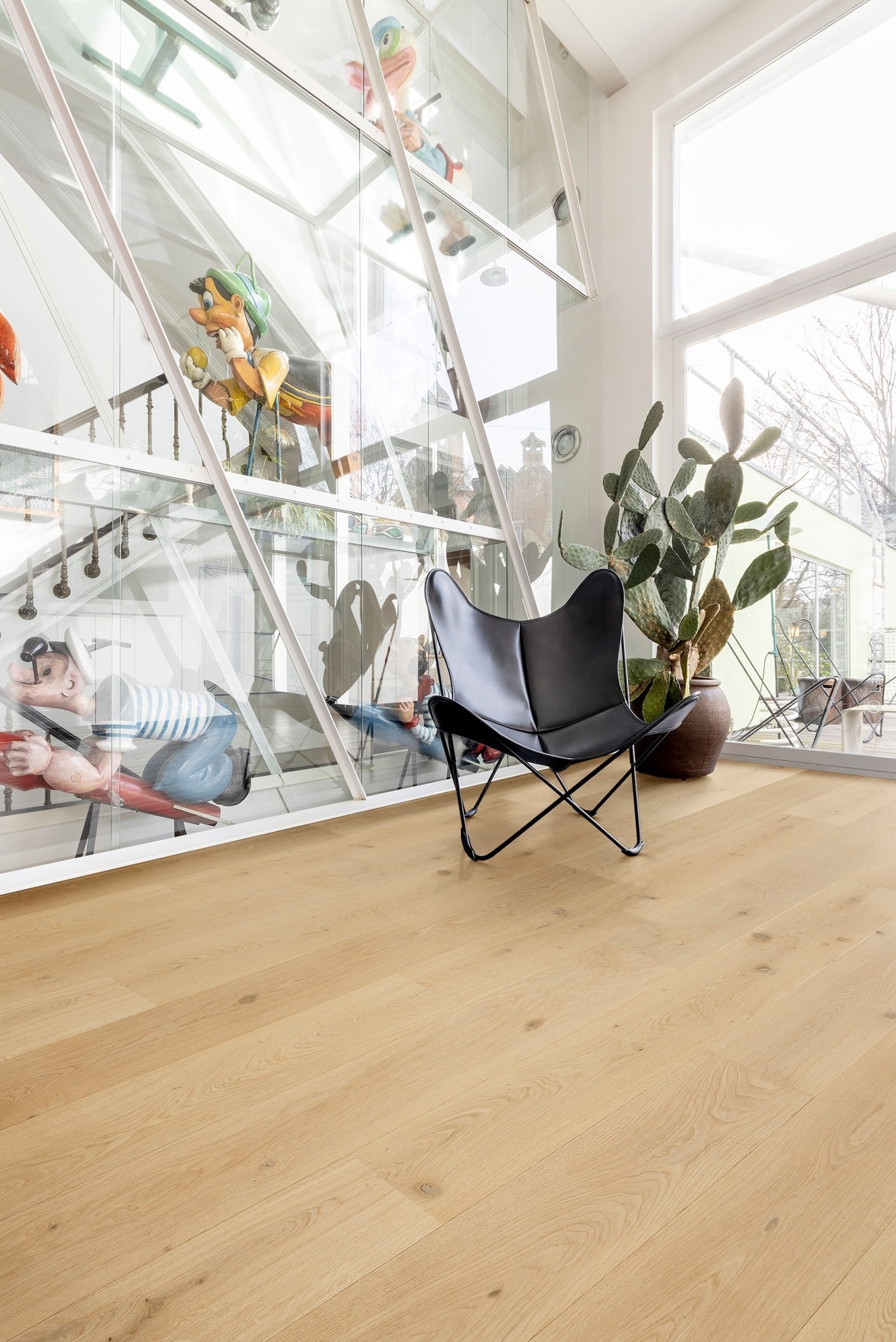 Parquet chêne clair dans un intérieur lumineux. Verrière architecturale exposant des statues de Pinocchio et Popeye. Fauteuil papillon et cactus.