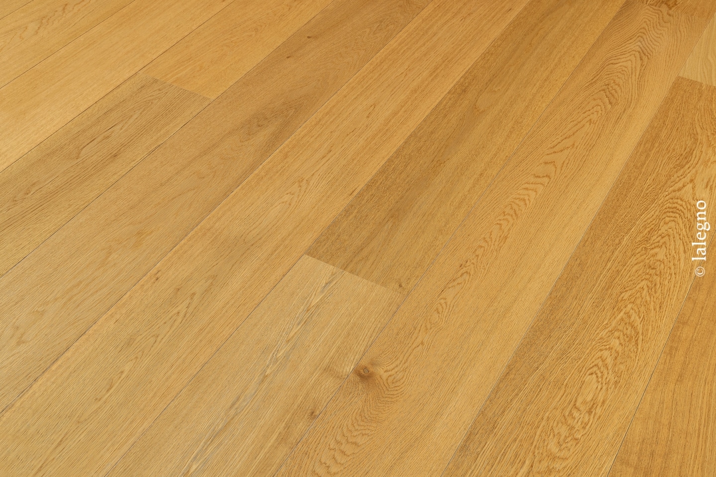 Détail d'un parquet en chêne clair aux lames larges et dorées, posé en angle. Grain du bois et petits nœuds visibles. Filigrane © lalegno.
