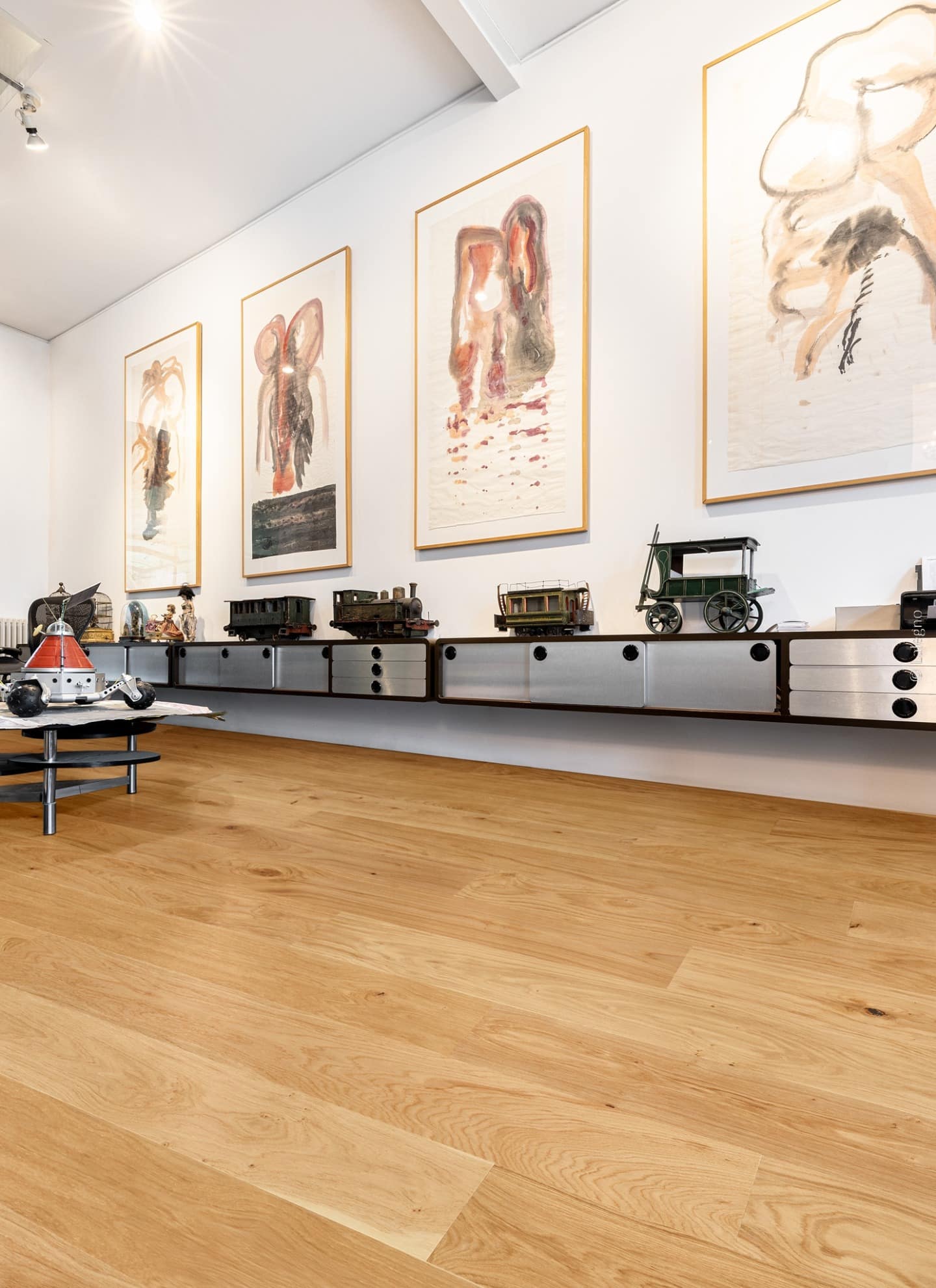 Parquet en chêne clair chaleureux dans une galerie. Meuble design flottant supportant des trains miniatures anciens et grands tableaux abstraits.
