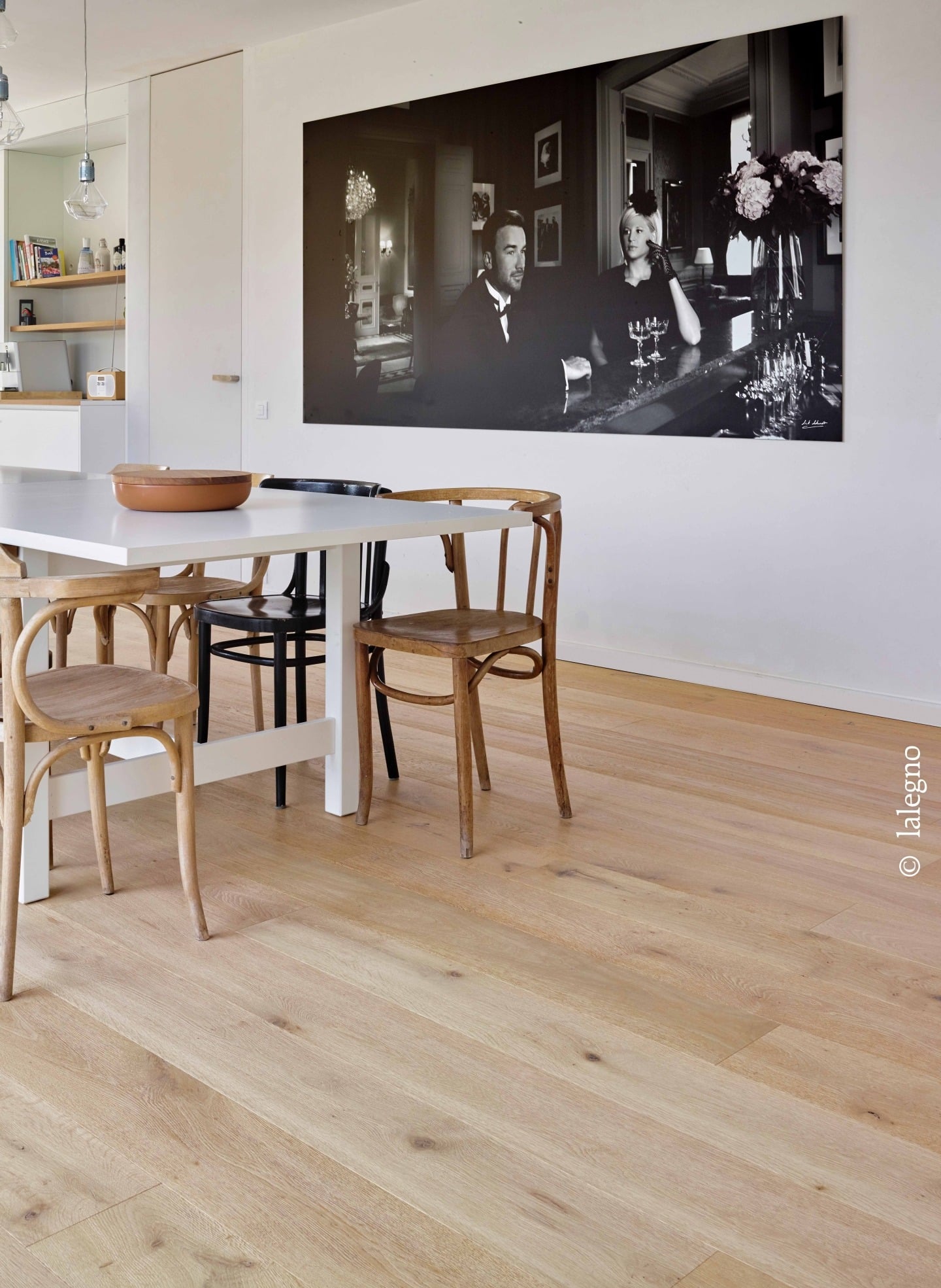 Parquet chêne clair à larges planches. Salle à manger moderne avec table blanche et chaises bistrot. Grande photo noir et blanc chic au mur.