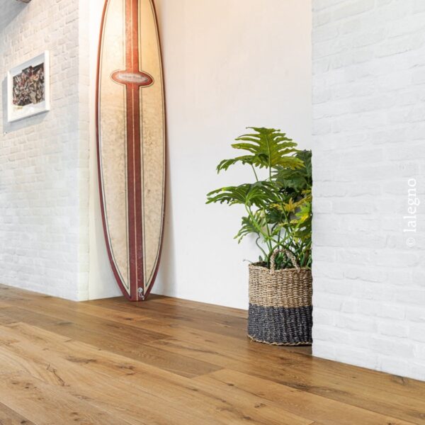 Parquet chêne miel. Décor intérieur lumineux avec murs en brique blanche, surfboard vintage et grande plante en panier tressé. © lalegno.