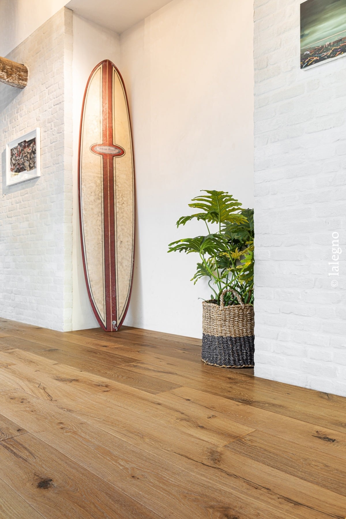 Parquet chêne miel. Décor intérieur lumineux avec murs en brique blanche, surfboard vintage et grande plante en panier tressé. © lalegno.