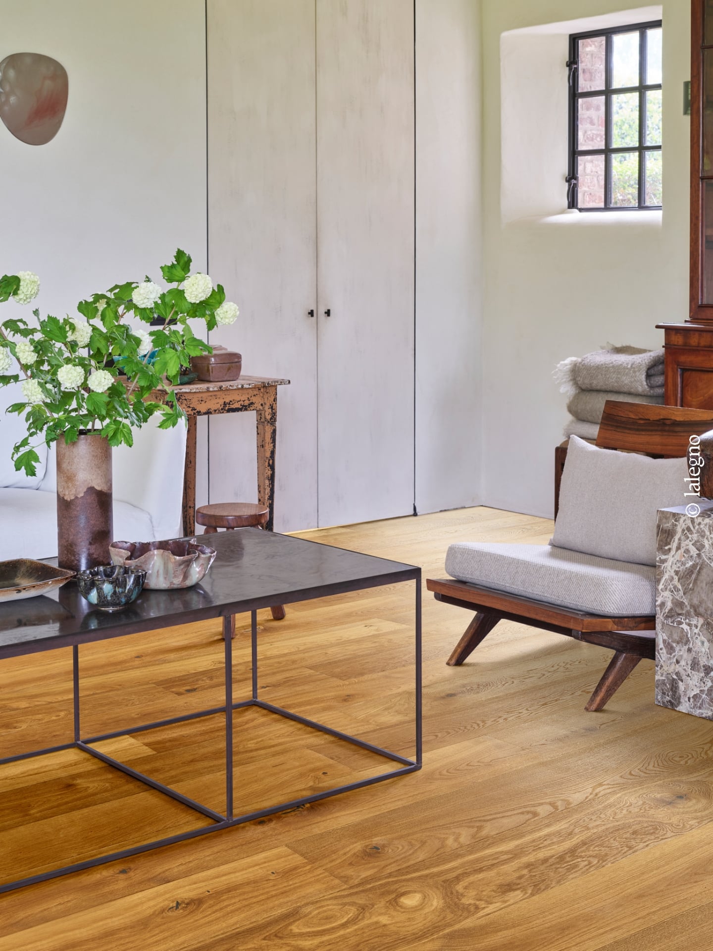 Parquet en chêne doré dans un salon rustique chic. Table basse en métal, fauteuil en bois, armoires patinées et fenêtre ancienne.