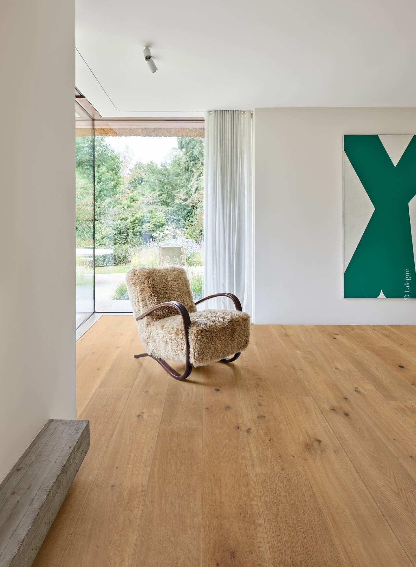 Fauteuil en fourrure devant une baie vitrée sur jardin. Plancher chêne naturel. Mur blanc, tableau vert abstrait.