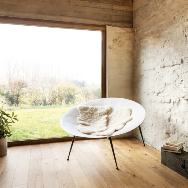 Fauteuil blanc design sur parquet chêne clair. Vue panoramique sur champ verdoyant. Mur pierre rustique et poutre en béton brut.