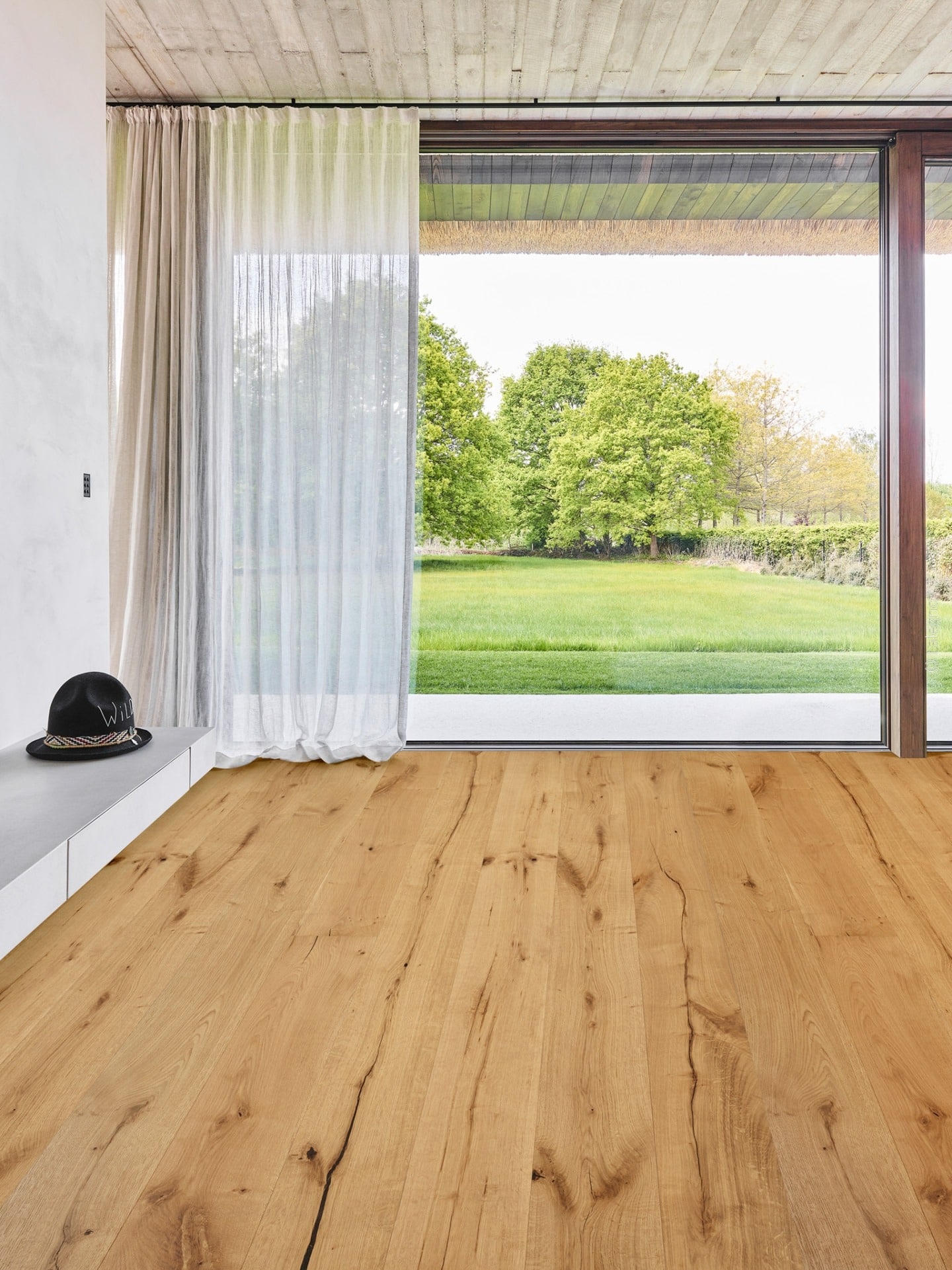 Parquet en chêne naturel rustique à lattes larges, fissures apparentes, baies vitrées et vue sur jardin. Plafond béton. Chapeau "Wild" sur console.