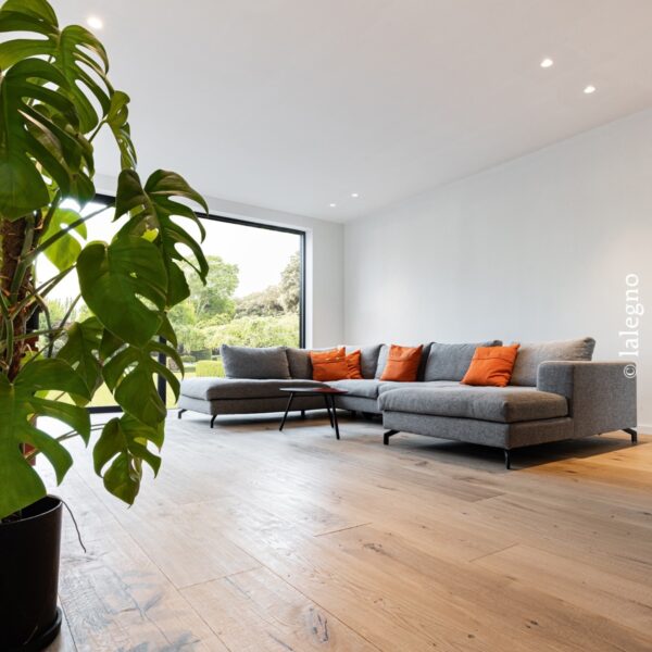 Salon moderne avec parquet en chêne clair. Grand canapé sectionnel gris et coussins orange, face à une large fenêtre et une plante Monstera.