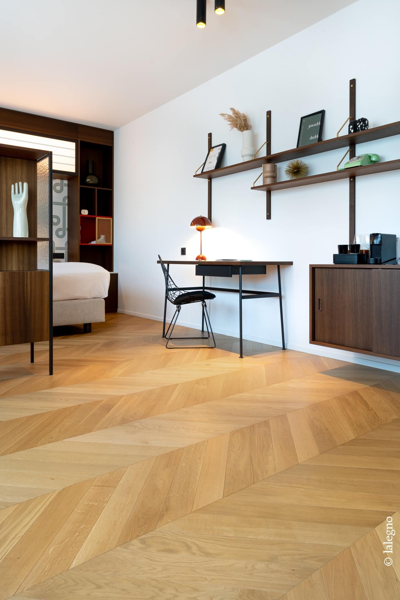 Parquet chevron en bois clair dans une chambre moderne. Étagères murales sombres, bureau, chaise design et séparation avec sculpture de main.