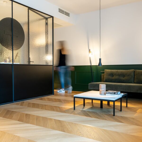 Salon moderne avec spectaculaire parquet chevron en bois clair. Cloison vitrée noire, canapé vert foncé et soubassement vert sapin.