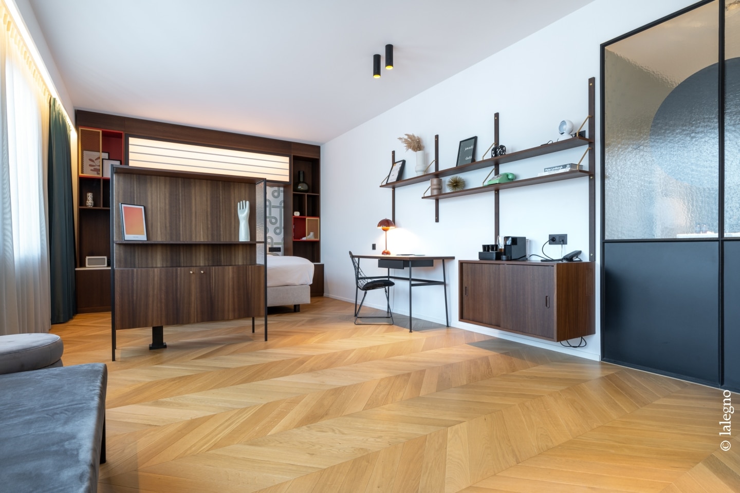 Suite moderne avec parquet chevron clair. Aménagement intérieur bois et blanc, séparateur de pièce, étagères murales design et coin bureau.