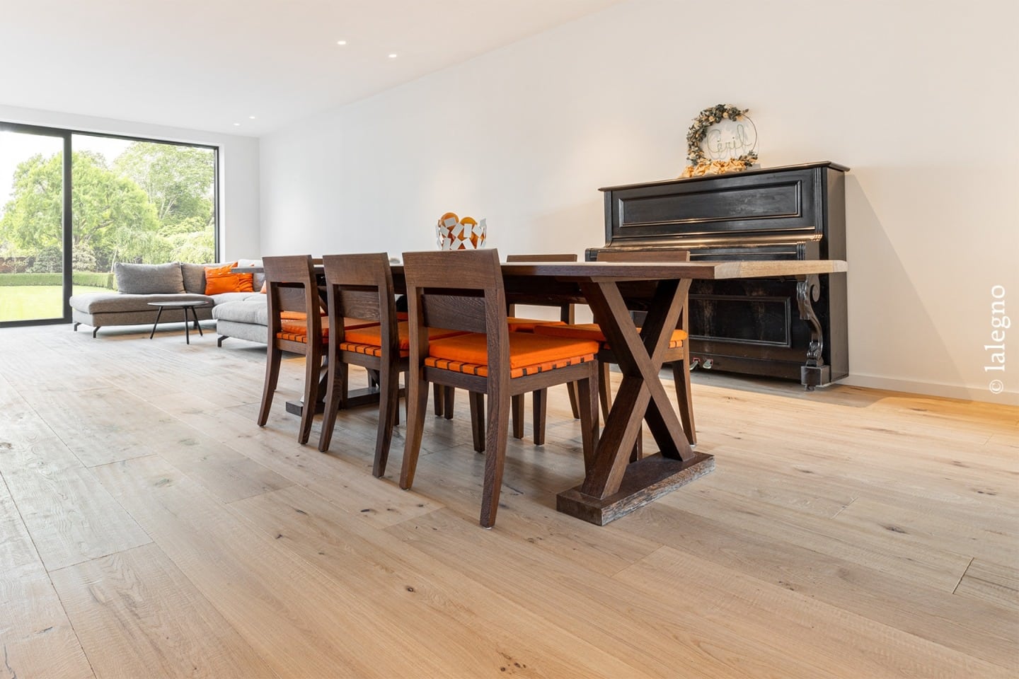 Salon/salle à manger moderne. Plancher chêne clair, mobilier bois foncé, coussins orange vifs. Piano et grande fenêtre sur verdure.