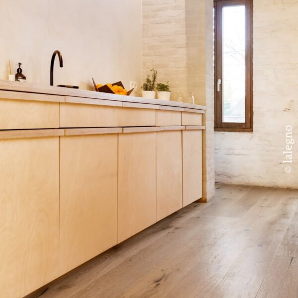 Cuisine contreplaqué bois clair, robinet noir, sur plancher en chêne. Murs de briques blanches et fenêtre. Poutre béton visible.