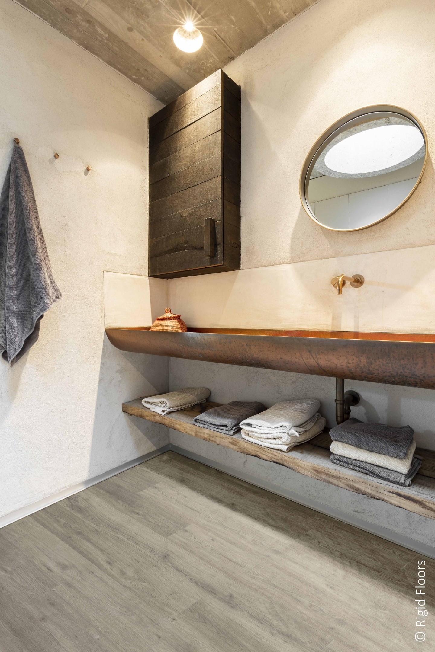 Salle de bain au design rustique industriel avec plancher rigide gris clair. Longue vasque en cuivre, miroir rond et armoire murale foncée.