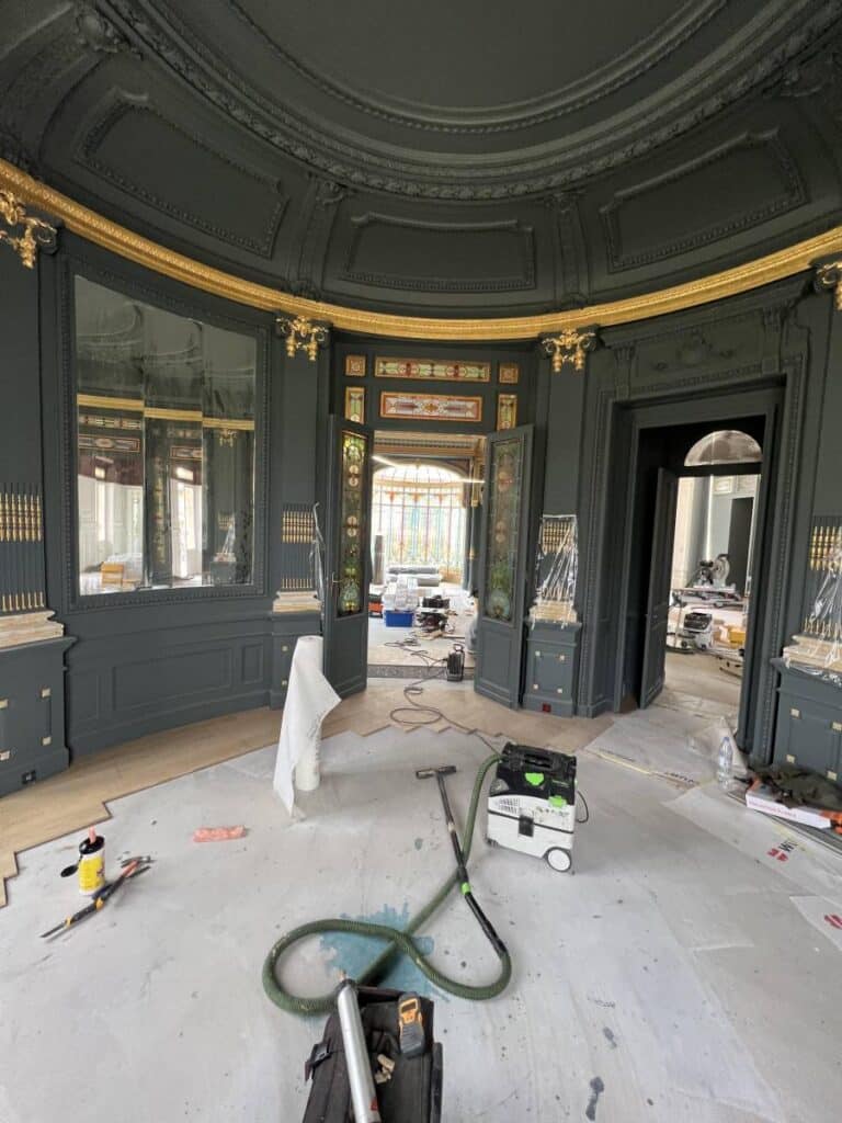 Rénovation luxe : Boiseries sombres, dorures et salon historique Salon luxueux en pleine rénovation. Boiseries vert-foncé, moulures et corniches dorées. Parquet en cours de pose, outils et aspirateur Festool au sol.