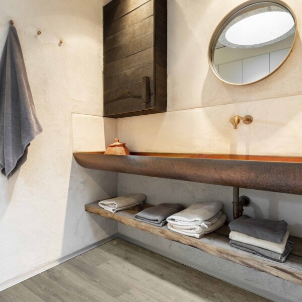 Salle de bain rustique. Sol vinyle effet bois clair, lavabo cuvette en cuivre martelé, meuble bois foncé et murs texturés.