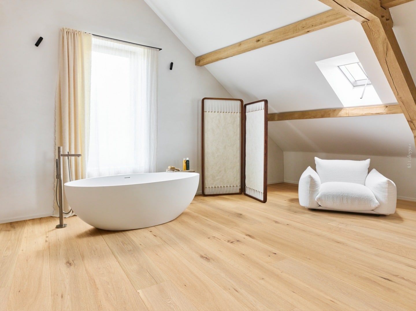 Luxueuse salle de bain sous-pente avec baignoire îlot blanche, parquet en chêne clair et charpente bois apparente.