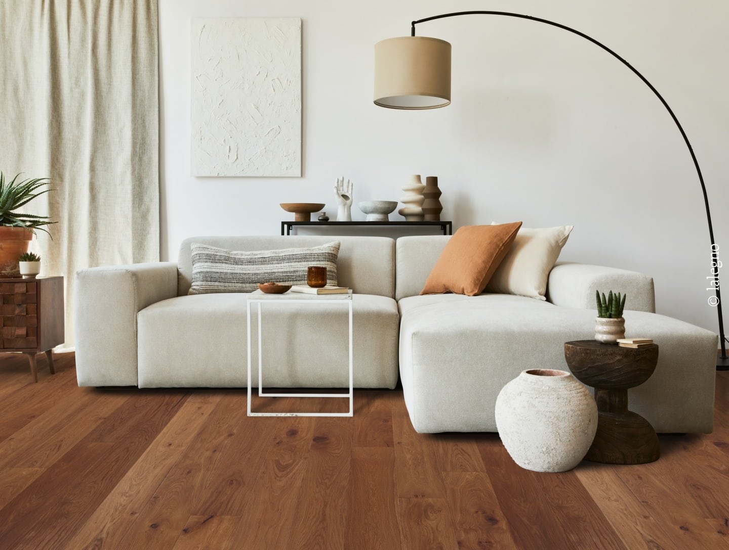 Salon minimaliste avec grand canapé modulaire beige, sol en parquet bois foncé, et décoration aux tons neutres terre cuite.