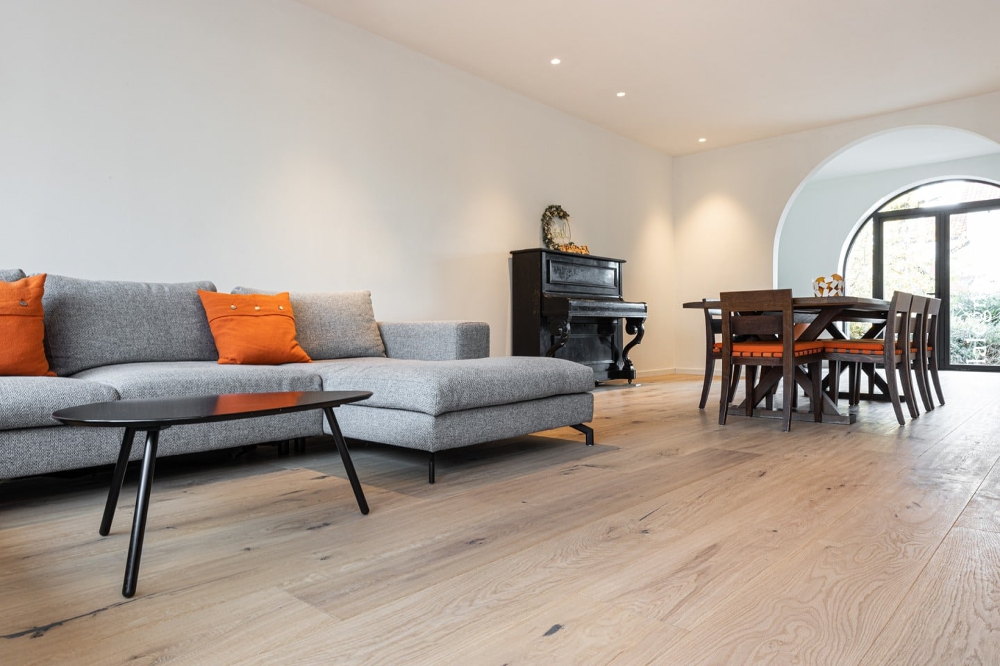 Salon ouvert moderne : sofa gris, accents orange, piano noir vintage et salle à manger. Parquet en chêne clair lumineux.
