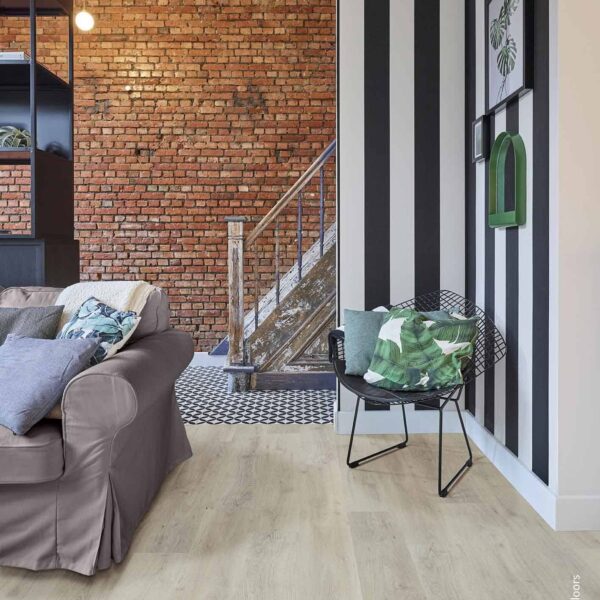 Salon industriel-chic: mur de briques, sol bois clair, et mur à rayures. Canapé gris, escalier rustique. © Rigid Floors.