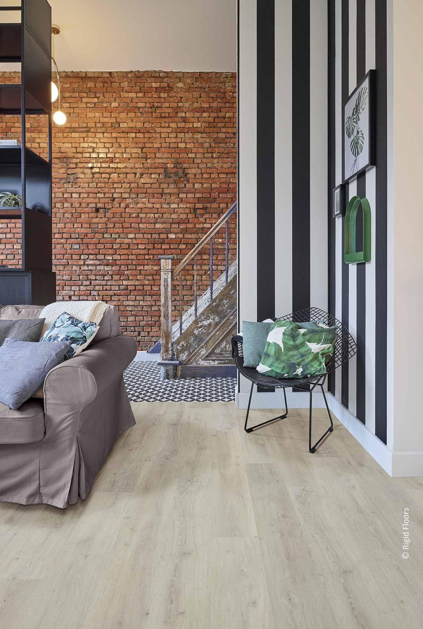 Salon industriel-chic: mur de briques, sol bois clair, et mur à rayures. Canapé gris, escalier rustique. © Rigid Floors.