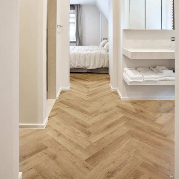 Couloir moderne avec sol rigide aspect chêne clair en chevrons. Vue sur une salle de bain blanche (lavabo) et une chambre lumineuse.