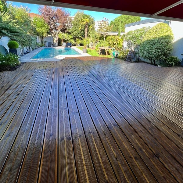 Grande terrasse en bois brun brillant menant à une piscine. Entretien d'une maison avec jardin luxuriant et palmiers ensoleillés.