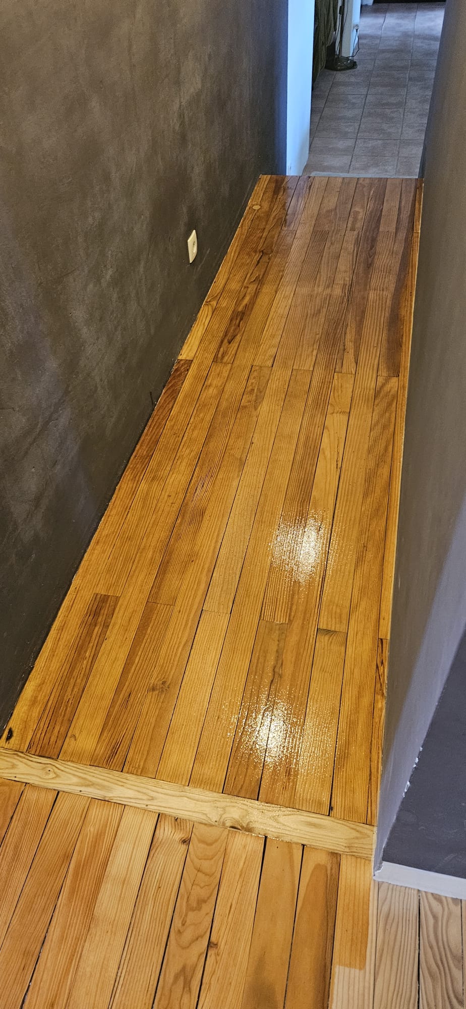 Couloir étroit avec parquet en bois clair vernis brillant et murs en béton ciré gris foncé.