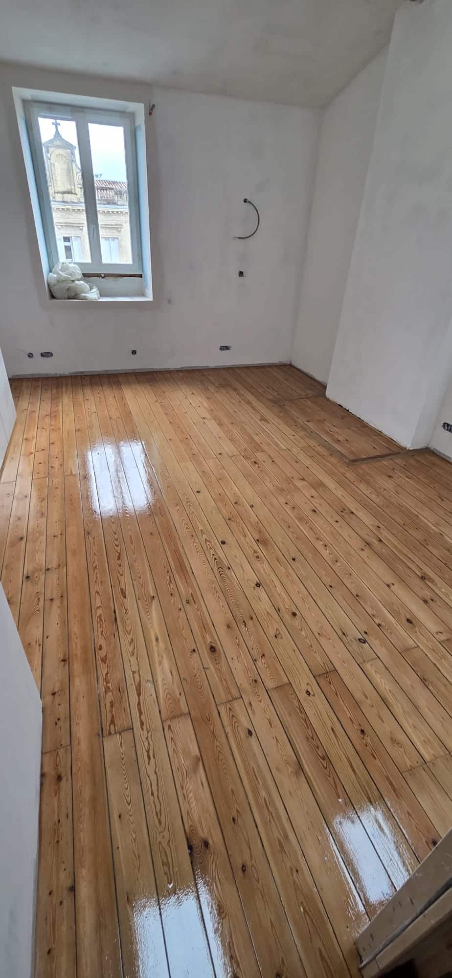 Parquet en bois massif poli dans une pièce lumineuse en rénovation avec murs blancs et vue sur une église.
