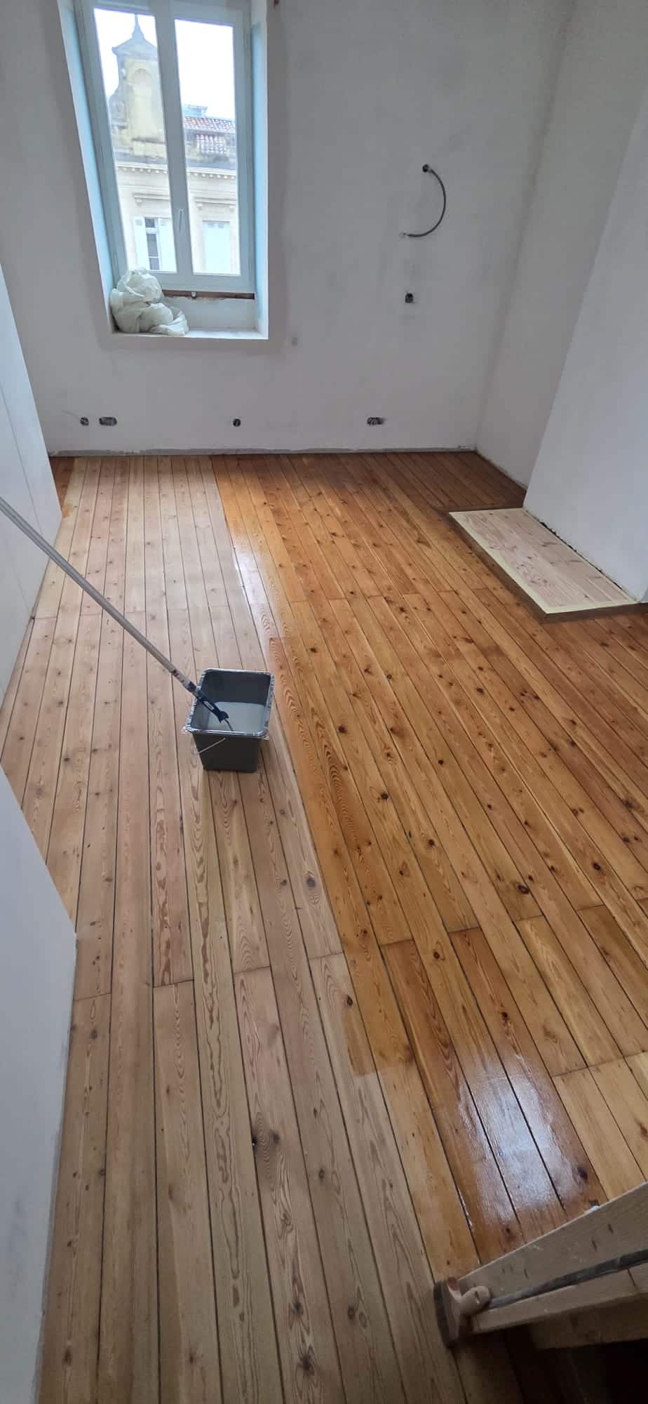 Vitrification d'un parquet en pin massif avec un rouleau dans une pièce lumineuse en rénovation.