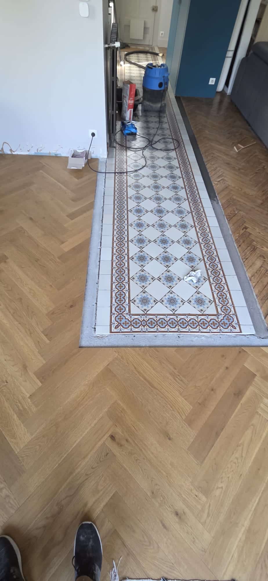 Rénovation de sol : Parquet chevrons et carreaux de ciment Parquet en chevrons entourant un tapis de carreaux de ciment à motifs bleus en cours de rénovation intérieure.