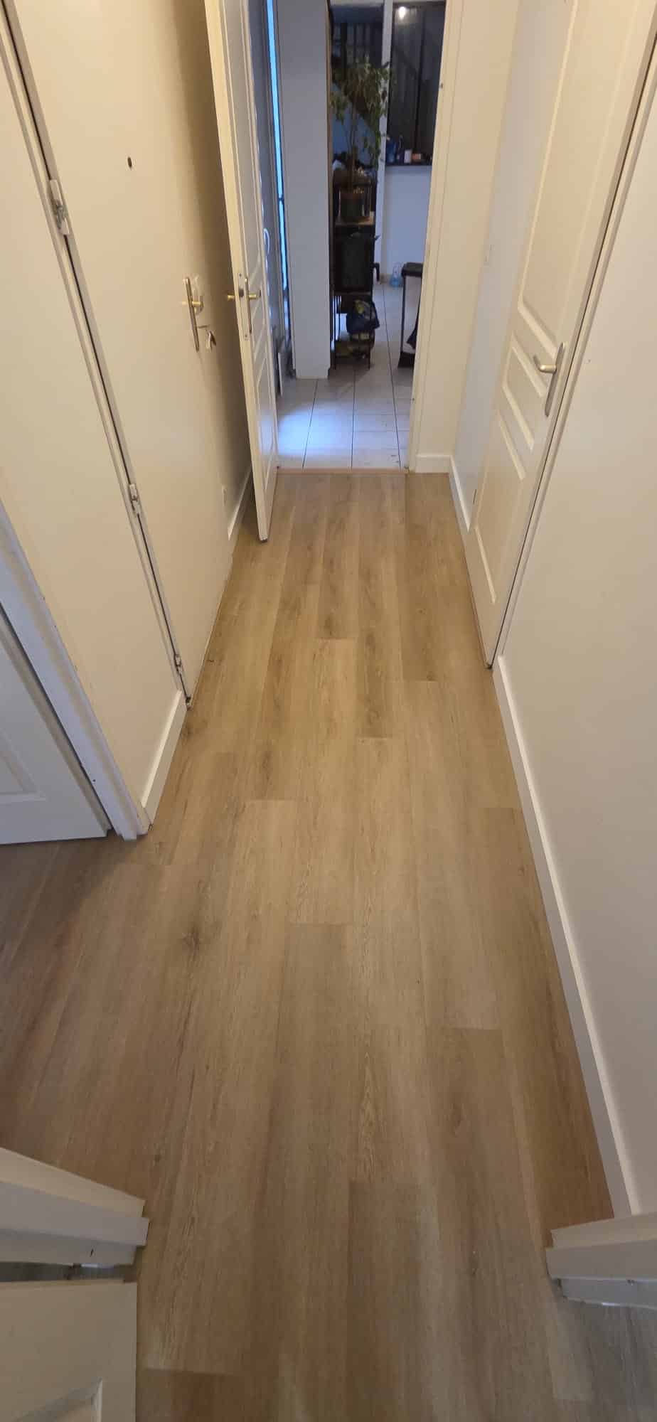 Couloir épuré avec sol en parquet de bois clair et murs blancs menant vers une pièce lumineuse.