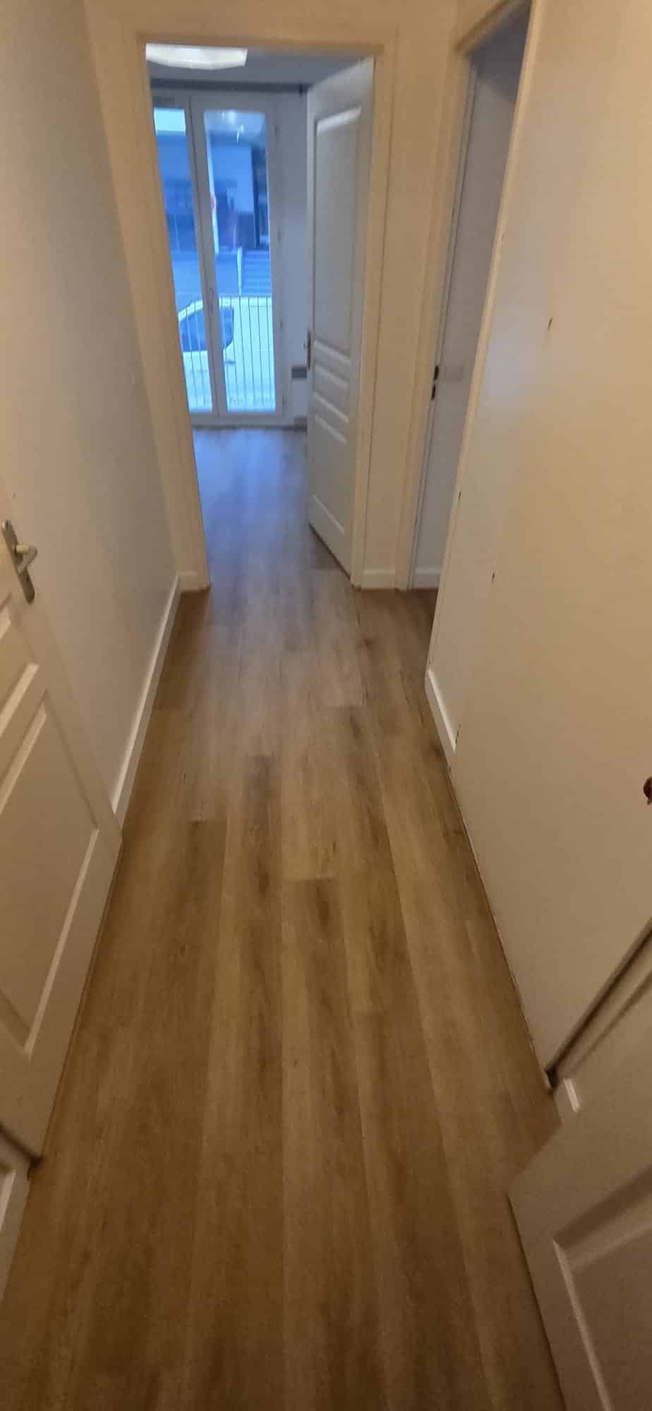 Couloir avec sol parquet bois clair, murs et portes blanches menant vers un balcon lumineux.