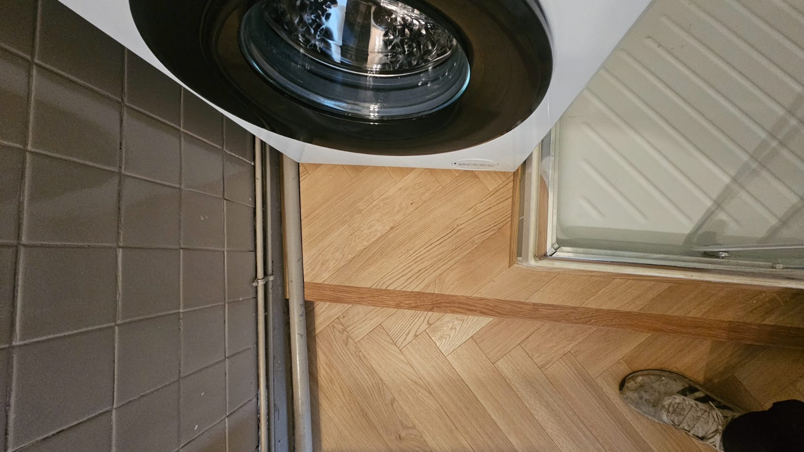 Lave-linge blanc sur sol en parquet chevron bois clair à côté d'un mur en carrelage gris et tuyauterie apparente.