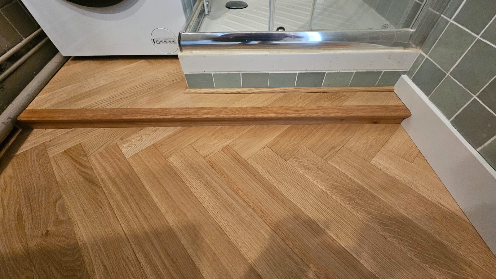 Parquet chêne chevrons dans une salle de bain avec barre de seuil en bois massif et carrelage mural vert.